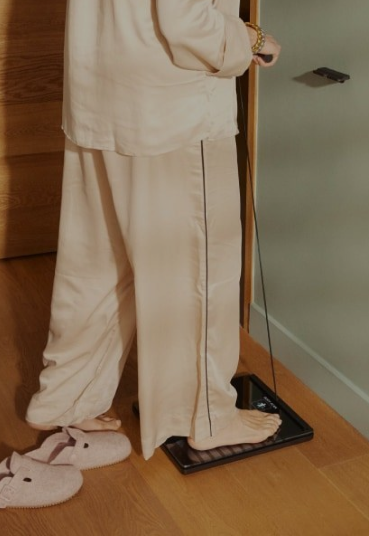 Person in neutral pyjamas standing on a Withings scale in a softly lit bedroom.